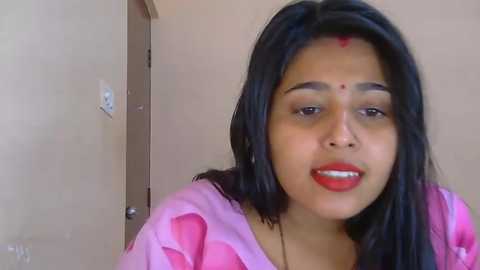 Video of a young South Asian woman with medium skin tone, long black hair, and a red bindi, wearing a pink saree. She smiles warmly in a plain indoor setting.