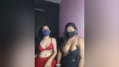 Video of two women with long dark hair, wearing matching blue masks, red and black lingerie, standing in a dimly lit room with black and pink walls.