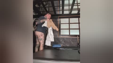 A video captures a tattooed, shirtless man in a black kilt carrying a white kimono in a traditional Japanese room with tatami mats and shoji screens.