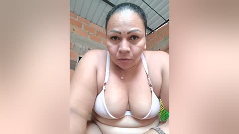Video of a plus-sized, light-skinned woman with dark hair pulled back, wearing a white bikini top, indoors with brick walls and corrugated metal ceiling.