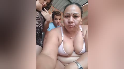 A video of a plus-sized woman in a white bra, with a young man and woman in the background. The woman has a neutral expression, and the man looks away.