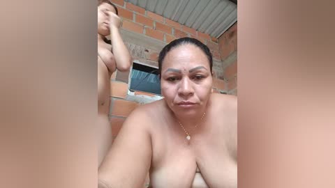 A video of a nude older woman with medium skin tone and dark hair, standing in a rustic outdoor area with brick walls and a corrugated metal roof.