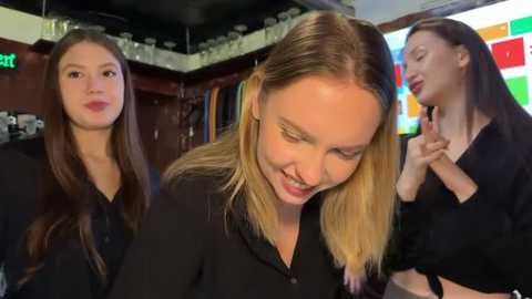Video of three women with long hair, wearing black, in a bar setting with colorful lights and glasses.