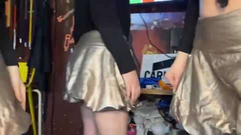 Video of two women in a cluttered room, one in a black top and metallic silver skirt, the other in a gold dress, holding a black garment.