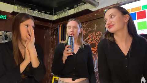 Video of three young women with fair skin and light brown hair, one drinking from a can, in a dimly lit bar with a colorful chalkboard wall.