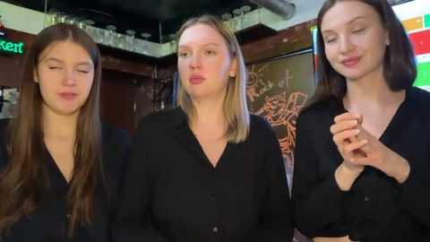 Video of three women with long hair, wearing black blouses, smiling and clapping in a dimly lit bar with colorful neon signs and shelves in the background.
