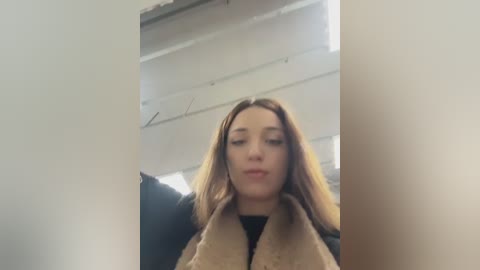 A video shows a young woman with straight, light brown hair, wearing a beige fur-trimmed coat, standing indoors against a white wall with a metal ceiling.