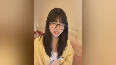 Video of an Asian woman with straight black hair, wearing glasses, a yellow cardigan, and a white shirt, smiling with her tongue sticking out, in a beige room with a bed and red object in the background.