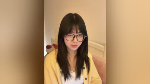 Video of an East Asian woman with long black hair, wearing glasses, a yellow cardigan, and a white top, smiling against a beige wall background.