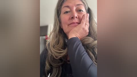 Video of a middle-aged woman with long, wavy brown hair, light skin, and a slight smile. She rests her chin on her hand, wearing a dark sweater. Background is blurred, indoors with neutral tones.