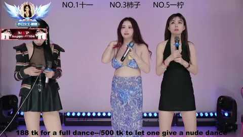 Video of three women singing into microphones, dressed in stylish, colorful outfits; one in a black bodysuit, one in a blue patterned dress, and one in a black dress.