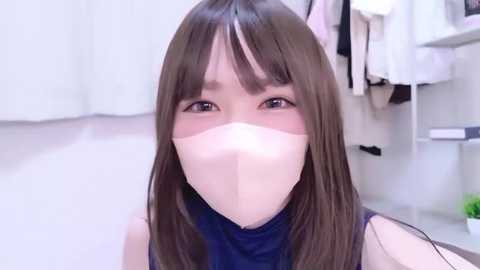 Video of a young Asian woman with long brown hair, wearing a blue sleeveless top, a white mask, and a soft, diffused glow. Background features a white bathroom with hanging towels and a green bottle.