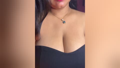 Video of a woman with medium brown skin, wearing a strapless black dress that accentuates her large breasts. She has long, dark hair and a dainty necklace with a small pendant. The background is out of focus.