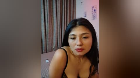 Video of an Asian woman with long black hair, medium skin tone, wearing a black bra, standing in a dimly lit room with beige walls, framed art, and a gray curtain.