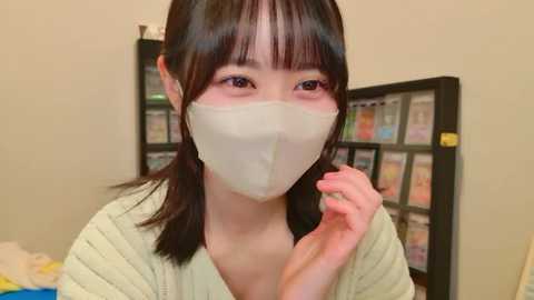 Video of an Asian woman with straight black hair and bangs, wearing a white face mask, beige sweater, and light makeup. Background shows a black bookshelf filled with colorful books and papers.
