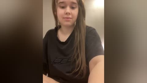 Video of a young woman with long brown hair in a black t-shirt, blurred background, looking down.