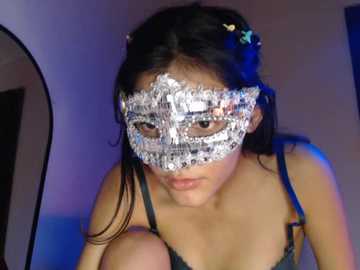 A video of an Asian woman with long black hair wearing a glittery masquerade mask, dark top, and blue background.