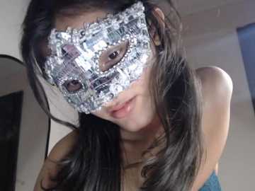 Video of an Asian woman with long, dark hair wearing a silver masquerade mask with intricate floral patterns. She has a light skin tone and is indoors, with a blurred background of neutral-colored walls.