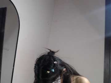 Video of a woman with long black hair, wearing a beige top, captured from behind, standing in a dimly lit room with white walls and a mirror.