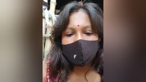 Video of an Indian woman with medium brown skin, wearing a black mask covering her nose and mouth, and a red bindi on her forehead. She has long, wavy black hair. Background shows blurred wooden structures.