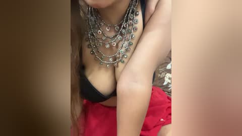 Video of a light-skinned woman with long hair, wearing a black bra, multiple silver necklaces, and red fabric, crouched in a dimly lit room.