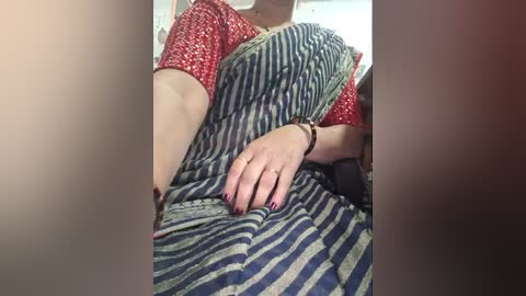 A video captures a woman in a red and white patterned saree, draped over a blue and white striped bedspread, with a ring-adorned hand resting on her lap. The background is blurred, with a soft, warm lighting.
