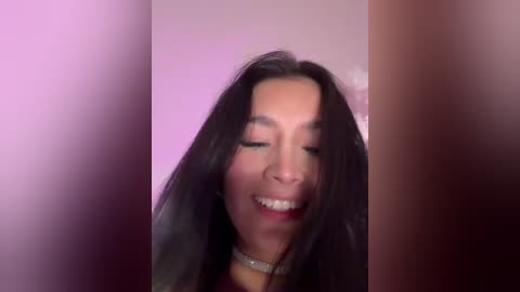 A video of a young woman with long, straight black hair, smiling widely, set against a blurred background with a soft pink and purple hue. She wears a silver choker necklace.