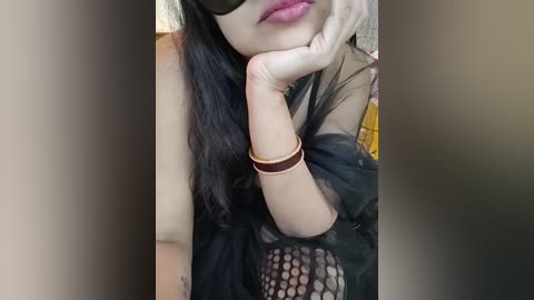 Video of a light-skinned woman with long black hair, wearing a black sheer mesh top, a brown bracelet, and a black mask, resting her chin on her hand.