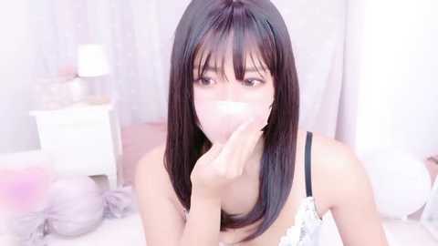 Video of an Asian woman with straight black hair, wearing a white lace bra, blowing into a tissue, in a pastel-themed bedroom.