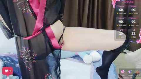 A video of a person in a sheer black robe with floral patterns, revealing pink lingerie and black panties, seated on a white surface. The background features a soft, light blue blanket.