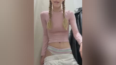Video of a young, slender woman with long, blonde braids, wearing a pink long-sleeve crop top and white, striped underwear, standing in a dimly lit dressing room.