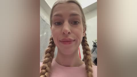 Video of a Caucasian woman with fair skin, light brown hair styled in twin braids, wearing a pink top. She has a neutral expression and is indoors, possibly in a bathroom.