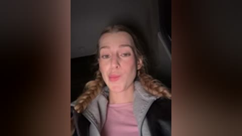 Video of a young woman with light skin, braided hair, and light pink shirt, seated in a car, wearing a black leather jacket.