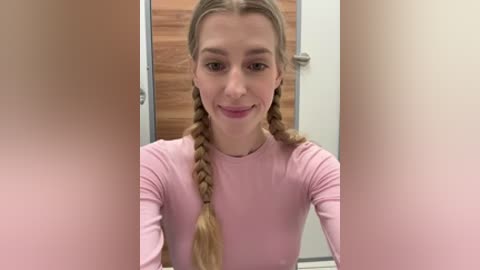 Video of a young Caucasian woman with light skin, long blonde braided pigtails, and a pink long-sleeved top. She is smiling and standing in front of a wooden door with a silver handle.