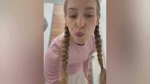 Video of a young, fair-skinned woman with blonde twin braids, wearing a light pink long-sleeved top and beige skirt. She is blowing a kiss from a narrow hallway with wooden floors and white walls.