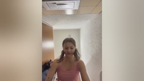 Video of a young woman with light skin, wearing a pink tank top, walking down a narrow, beige-tiled hallway with textured white walls.