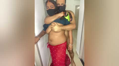 A candid video of a shirtless, muscular man with a black mask, embracing a topless woman in a red saree with a black mask, in a narrow hallway.