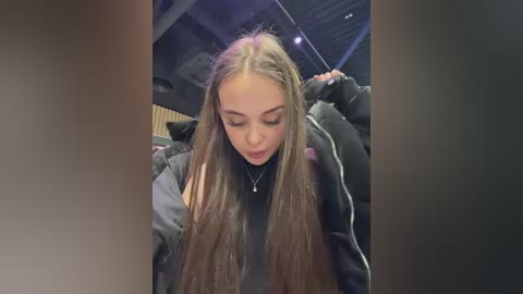 Video of a young woman with long, straight brown hair, wearing a black jacket, standing in a dimly lit room with metallic ceiling panels and string lights.