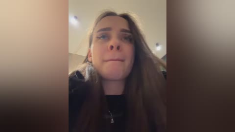 Media: Video of a young woman with long brown hair, wearing a black jacket, with a neutral expression, taken indoors with dim lighting.