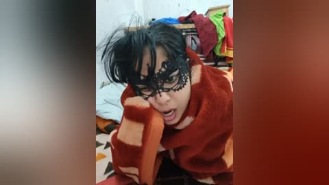 A video shows a young person with short black hair, wearing a black lace mask, a red and orange striped blanket, and a distressed expression, kneeling on a colorful patterned rug in a cluttered room.