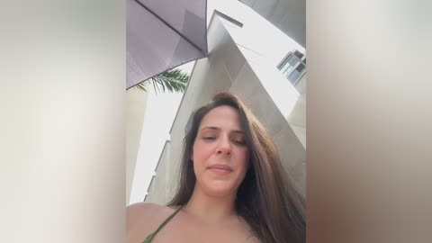 Video of a young woman with long brown hair, wearing a green bikini, standing in a modern, minimalist outdoor setting with white buildings and a palm tree in the background.