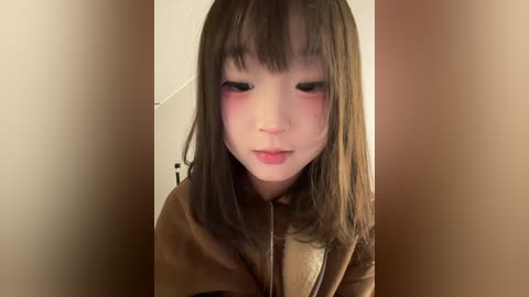 Video of a young Asian woman with long, straight brown hair, wearing a brown jacket, with flushed cheeks and a neutral expression. Background is blurred, featuring a beige wall and a corner.