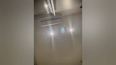 Video of a modern, beige-walled room with a central ceiling-mounted air conditioner, three bright, blinding lights, and a partially visible rectangular vent.
