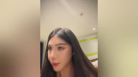 A video of a young woman with long, straight, dark hair, fair skin, and red lipstick, smiling slightly. The background features a blurred, modern interior with green walls and a light-colored ceiling.