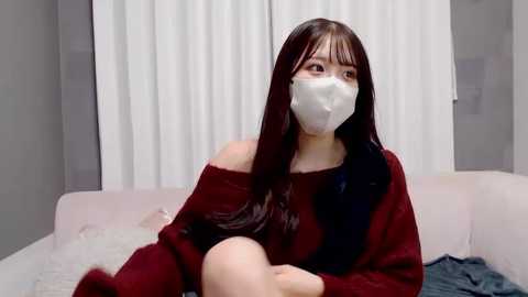 A video of a young East Asian woman with long, dark hair, wearing a red sweater and white face mask, sitting on a light pink sofa in a modern, minimalistic living room with white curtains.