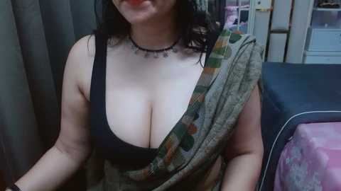 Video of a plus-sized woman with dark hair, wearing a low-cut black tank top revealing cleavage, draped in a patterned sarong, sitting in a dimly lit room with shelves and a bed in the background.