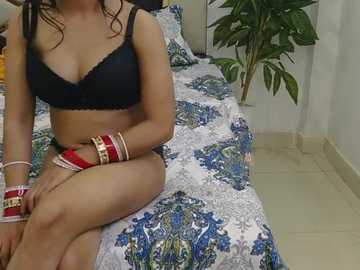 Video of a woman with medium skin tone, wearing black lingerie and colorful bangles, sitting on a bed with blue and white paisley-patterned sheets, green potted plant in the background.