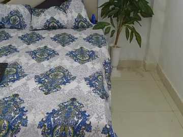 Video of a neatly made bed with a white and blue paisley-patterned comforter and pillowcases. The room has a beige tiled floor, a potted plant in the corner, and a plain white wall.