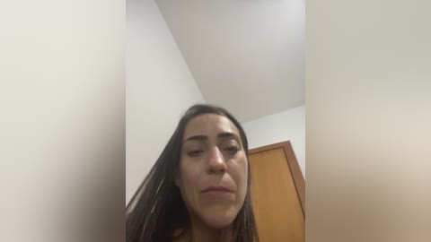 Video of a woman with long dark hair, fair skin, and closed eyes, making a pouty face, standing in a small, white-walled room with a wooden door and ceiling fan.