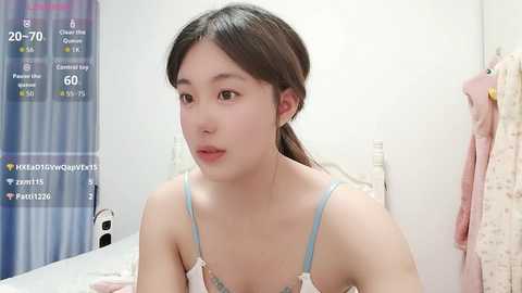 A video of a young Asian woman with fair skin, long black hair, and a light blue spaghetti strap top, sitting on a bed with pink and white bedding. The background features a plaid blanket and a digital clock displaying 20:17.